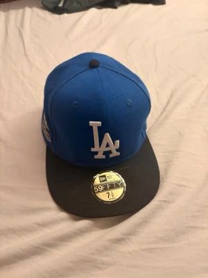 Blå LA Dodgers keps New Era 59FIFTY - Snygg blå keps från New Era 59FIFTY med svart skärm och LA Dodgers-logga framtill. På sidan finns ett broderat World Series-märke. Klassisk flat brim och strukturerad passform, perfekt för dig som gillar streetwear och sportig stil.