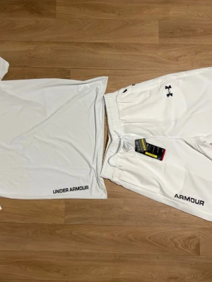 Vitt träningsset från Under Armour - Säljer ett vitt träningsset från Under Armour med t-shirt och shorts. Båda plaggen har logga och textdetaljer i svart. Shortsen har resårmidja med dragsko och sidofickor. Perfekt för gym eller löpning.