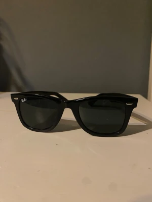 Svarta Ray-Ban Wayfarer solglasögon - Klassiska svarta Wayfarer solglasögon från Ray-Ban med bred båge och mörka glas. Snygg retrostil med Ray-Ban-logga på linsen och sidan. Passar perfekt till alla outfits och ger en cool vibe.