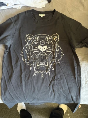 Svart Kenzo t-shirt med tigertryck - Snygg svart t-shirt från Kenzo med stort tigertryck och texten 'Paris' framtill. Klassisk rund halsringning och korta ärmar. Tillverkad i mjuk bomull som känns skön mot huden. Perfekt för dig som vill sticka ut med en ikonisk design.