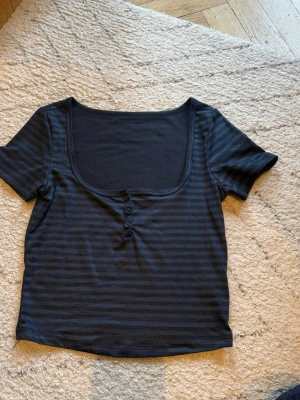 Marinblå randig topp med knappar - Kortärmad svart topp med diskreta ränder och djup rundringning. Tre knappar framtill ger en snygg detalj. Perfekt till jeans eller kjol för en enkel men stilren look.