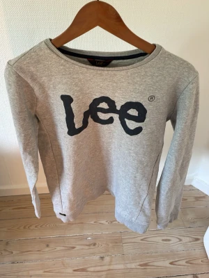 Grå sweatshirt från Lee - Snygg grå sweatshirt från Lee med stort svart tryck på bröstet. Tröjan har rund halsringning, långa ärmar och ribbade muddar vid ärmslut och nederkant. Perfekt för en avslappnad och trendig stil.
