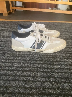 Vita och beige sneakers från Pepe Jeans - Snygga sneakers från Pepe Jeans i vitt och beige med mörkblå detaljer och två ränder på sidan. Skorna har klassisk låg profil, vita snören och en slitstark sula. Materialmix av mocka och textil ger en fräsch och sportig look.