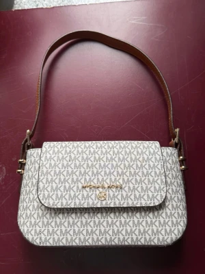 Michael Kors beige axelremsväska - Snygg beige handväska från Michael Kors med MK-logga över hela väskan. Väskan har guldfärgade detaljer, justerbar brun axelrem och en praktisk ficka med dragkedja på baksidan. Perfekt storlek för det viktigaste och stängs med lock framtill.