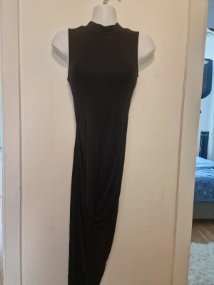 Svart ärmlös långklänning - Svart klänning med hög krage en draperad detalj framtill som skapar en asymmetrisk fåll.