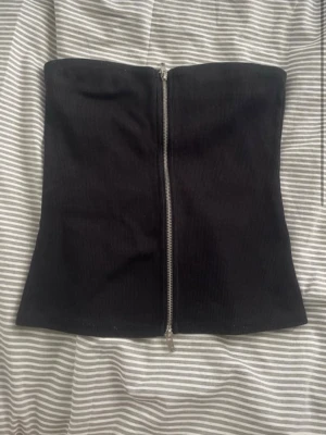 Svart tubtopp med dragkedja - Säljer en svart tubtopp i ribbat material med en snygg dragkedja framtill. Toppen är axelbandslös och har en tight passform som framhäver figuren. Perfekt att styla med jeans eller kjol för en trendig look.