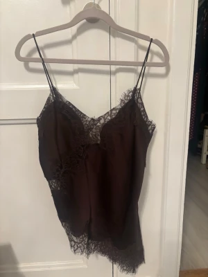 Viral Zara topp - Mörkbrun linnetopp med tunna axelband och vacker spets längs urringning och nederkant. Toppen har en mjuk, glansig finish och ger en elegant känsla med sina fina spetsdetaljer. Perfekt att styla lager på lager eller som den är.