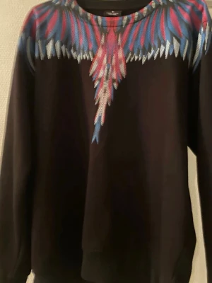 Marcelo burlon  - Cool svart hoodie med ett färgglatt fjädermönster i rött, blått och vitt över axlar och bröst. Tröjan har rund halsringning och långa ärmar. Perfekt för dig som vill sticka ut med en unik design.