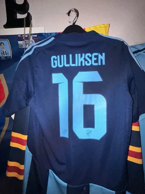Djurgården matchtröja Gulliksen #16 - Säljer en Djurgården matchtröja från Adidas med namn Gulliksen och nummer 16 på ryggen. Tröjan är blå med ljusblå ränder, klubbmärke på bröstet och sponsortryck. Korta ärmar med färgglada detaljer och klassisk sportig passform. Perfekt för fotbollsfans!