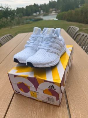 Vita Adidas Ultraboost sneakers - Fräscha helvita Adidas Ultraboost sneakers med stickad ovandel och klassiska tre ränder på sidan. Skorna har en bekväm Boost-sula och svart yttersula. Perfekta för dig som gillar stilrena och sportiga sneakers med grym komfort.
