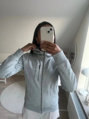 Sail Racing hoodie  - Snygg grå hoodie från Sail Racing med dragkedja hela vägen, justerbar huva och en bröstficka med dragkedja. Tillverkad i mjukt material som är perfekt för chill eller träning. Diskret logga på bröstet och stilren design.