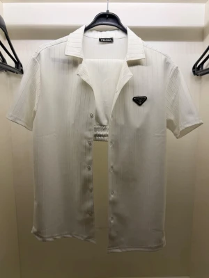 PRADA overshirt + shorts set - Säljer en stilren vit skjorta från Prada med korta ärmar och diskreta vertikala ränder. Skjortan har klassisk krage, knappar framtill och en svart Prada-logga på bröstet. Perfekt för dig som gillar minimalistisk och exklusiv design.