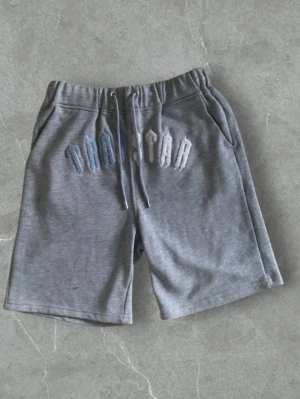Grå shorts från Trapstar med snörning - Grå shorts från Trapstar med broderad logga framtill i olika nyanser av blått och vitt. Modellen har elastisk midja med snörning och två sidofickor. Perfekta för sommaren och riktigt sköna att bära till vardags.