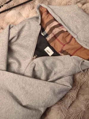 Grå hoodie med dragkedja från Burberry  - Snygg grå hoodie med dragkedja och klassisk känguruficka. Insidan av huvan har ett rutigt mönster i beige, brunt och rött som ger en unik touch. Tillverkad i mjukt sweatshirtmaterial och har justerbar dragsko i huvan. Perfekt för en chill och avslappnad stil.