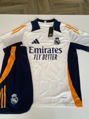 Real Madrid matchställ Adidas S - Säljer ett Real Madrid matchställ från Adidas i storlek S. Setet består av en vit tröja med mörkblå och orange detaljer samt matchande shorts. Tröjan har klubbmärke, Adidas-logga och 'Emirates Fly Better' tryckt framtill. Materialet är lätt och andas, perfekt för fotboll.