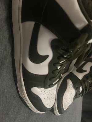Svarta och vita höga sneakers - Snygga höga sneakers i svart och vitt med klassisk design och perforerad tåbox. Skorna har en tydlig swoosh-detalj på sidan och snörning hela vägen upp. Materialet är en mix av slätt läder och textil, vilket ger en stilren och sportig look.