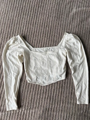 Vit långärmad korsettopp - Trendig vit korsettopp med långa puffärmar och markerade sömmar framtill. Toppen har en croppad passform och fyrkantig urringning som ger en snygg siluett. Perfekt att styla med höga jeans eller kjol för en cool look.