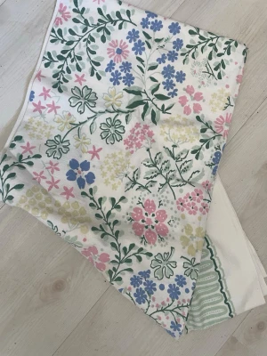 Blommigt tygstycke i pastellfärger - Säljer en Tetro Fuk ca 75.75 i mått med blommigt mönster i pastellfärger som rosa, blått, grönt och gult på vit botten. Tyget har en mjuk känsla och passar perfekt för DIY-projekt eller som accessoar. Mönstret är livligt med olika sorters blommor och blad.