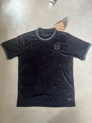 Svart Brasilien fotbollströja Nike - Snygg svart fotbollströja från Nike med Brasilien-emblem på bröstet. Tröjan har rund hals med grå och vita detaljer samt korta ärmar. Tillverkad i ett lätt och ventilerande material som passar perfekt till träning eller match.