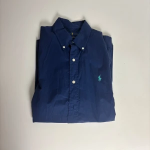 Blå skjorta från Ralph Lauren Slim Fit - Snygg marinblå skjorta från Ralph Lauren i slim fit-modell. Skjortan är i 100% bomull med stretch och har klassisk krage, vita knappar och broderad logga på bröstet. Perfekt för dig som gillar stilrena och tidlösa plagg.
