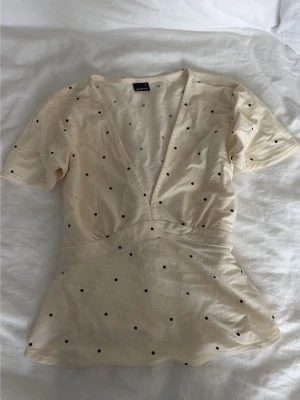 Prickig omlottopp med knytband - Söt ljusbeige omlottopp med svarta prickar och djup v-ringning. 