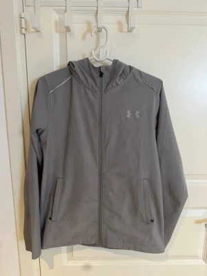 Grå vindjacka från Under Armour - Snygg grå vindjacka från Under Armour med dragkedja framtill, huva och två fickor med dragkedja. Jackan har meshfoder för extra ventilation och en stilren logga på bröstet. Perfekt för blåsiga dagar och sportiga outfits.