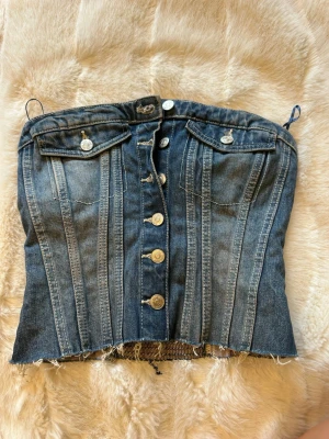 Jeans korsettopp med knappar - Snygg korsettopp i blå denim med markerade sömmar och silverfärgade knappar framtill. Toppen har två bröstfickor med lock och rå kant nertill för en edgy look. Axelbandslös design som ger en cool och trendig vibe.