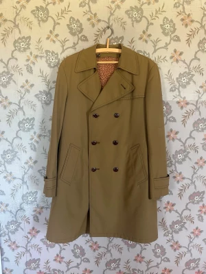 Klassisk beige trenchcoat med knappar - Stilren trenchcoat i beige med dubbelknäppning och bred krage. Jackan har två snedställda fickor och dekorativa manschetter vid ärmsluten. Tillverkad i ett slätt tyg som ger en tidlös look. Perfekt för dig som vill ha en klassisk och snygg jacka. Den är 100 cm lång 