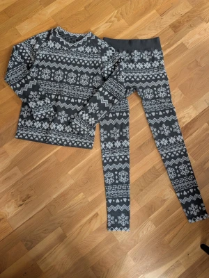 Mörkgrått underställsset med snöflingor - Säljer ett tvådelat underställsset i mörkgrått med vita snöflingor och geometriska mönster. Setet består av en långärmad tröja och matchande tights. Materialet ser ut att vara mjukt och stretchigt, perfekt för kalla dagar.