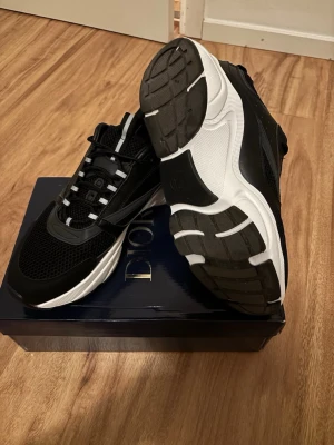 Svarta och vita sneakers med meshdetaljer B22 - Snygga svarta sneakers med vita detaljer och robust sula. Ovandelen är i mesh och syntetmaterial för en sportig look. Skorna har svarta och vita snören samt dragflik bak. Perfekta för dig som gillar stilrena och moderna sneakers.