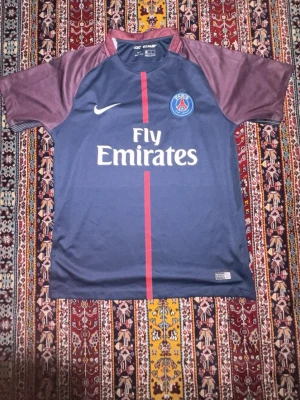 PSG matchtröja blå/röd Nike - Säljer en Paris Saint-Germain matchtröja från Nike i blått med vinröda ärmar och röd rand på framsidan. Tröjan har PSG-logga, Nike Swoosh och Fly Emirates-tryck. Tillverkad i Dri-FIT material som andas, perfekt för fotboll eller träning.
