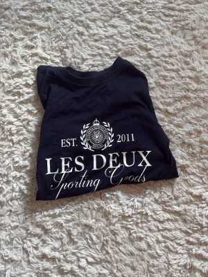 Svart Les Deux t-shirt med tryck - Snygg svart t-shirt från Les Deux med vitt tryck framtill där det står 'LES DEUX Sporting Goods' och 'EST. 2011'. Klassisk rund hals och korta ärmar. Perfekt till jeans eller shorts för en clean och stilren look.