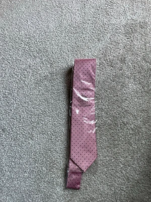 Rosa slips med små prickar - Snygg rosa slips med små svarta prickar. Slipsen har en klassisk form och är tillverkad i ett glansigt tyg som ger en elegant känsla. Perfekt accessoar för att lyfta din outfit med lite färg och stil.