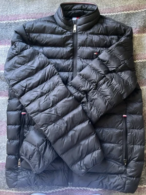 Svart dunjacka från Tommy Hilfiger - Snygg svart dunjacka från Tommy Hilfiger med quiltad design och dragkedja framtill. Jackan har ståkrage, två sidofickor med dragkedja och diskret logga på bröstet. Perfekt för kyliga dagar och enkel att matcha med olika outfits. Strl S | passar 175 till 180 utan problem 