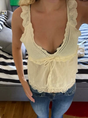 Söt Zara topp - Romantisk linnetopp i ljus beige med spetsdetaljer och volangkantade axelband. Djup v-ringning och knyt framtill ger en feminin look. Toppen är ärmlös och har broderade detaljer samt en luftig passform, perfekt för varma dagar.