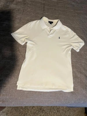 Vit pikétröja från Polo Ralph Lauren - Klassisk vit pikétröja från Polo Ralph Lauren med korta ärmar och krage. Tröjan har den ikoniska lilla logotypen broderad på bröstet och är tillverkad i mjuk bomull. Perfekt till jeans eller shorts för en clean och stilren look. Sitter som en Large!