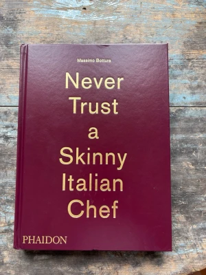 Never Trust a Skinny Italian Chef - En snygg bok med vinrött hårdband och guldfärgad text på omslaget. Skriven av Massimo Bottura och fylld med inspirerande bilder och berättelser om italiensk matlagning. Perfekt för dig som älskar mat och vill ha en unik bok i bokhyllan. Aldrig läst, helt oanvänd.