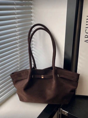 Mörkbrun tote handväska i mocka - Stilren mörkbrun handväska i mjuk mocka med långa dubbla axelband och minimalistisk design. Väskan har en enkel dekorativ rem framtill och rymlig insida, perfekt för dig som gillar klassiska och tidlösa accessoarer.