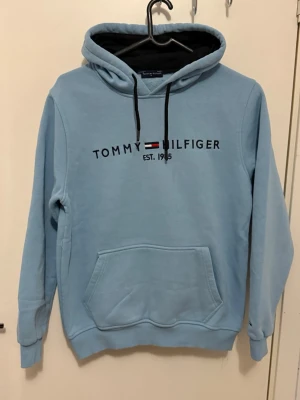 Ljusblå hoodie Tommy Hilfiger - Ljusblå hoodie från Tommy Hilfiger med svart dragsko i huvan och klassisk logga broderad på bröstet. Stor magficka framtill och ribbade muddar vid ärmar och nederkant. Perfekt för en avslappnad och stilren look.