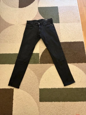Svarta slim fit jeans från Replay - Säljer ett par svarta slim fit jeans från Replay med klassisk femficksmodell och knappgylf. Jeansen har en stilren look och är tillverkade i ett mjukt denimtyg med lite stretch för extra komfort. Perfekta för dig som gillar en smalare passform.