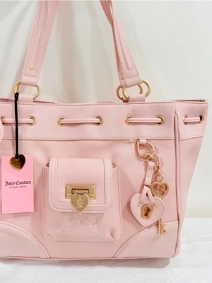 Ljusrosa handväska Juicy Couture - Supersöt ljusrosa handväska från Juicy Couture med guldfärgade detaljer, hjärtformade hängen och en liten ficka framtill med lås. Väskan har dubbla handtag, dekorativ rosett och klassisk logga. Perfekt för dig som gillar en feminin och trendig stil.