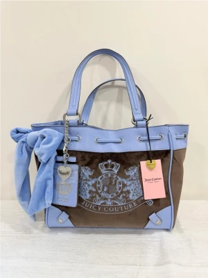 Juicy Couture brun-blå handväska daydreamer  - Snygg handväska från Juicy Couture i brun velour med ljusblå detaljer och handtag. Väskan har ett stort blått sidenband, silverfärgade metalldetaljer och ett emblem framtill. Perfekt för dig som gillar statement-accessoarer och vill sticka ut.
