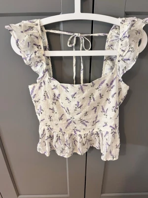 Blommig volangtopp från H&M - Supersöt topp från H&M i vitt med lila blommönster. Toppen har volangdetaljer vid axlarna, knytband i ryggen och smockad rygg för extra komfort. Perfekt till sommaren och ger en romantisk vibe till din outfit.