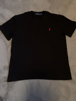Svart t-shirt från Polo Ralph Lauren - Klassisk svart t-shirt från Polo Ralph Lauren med röd broderad logga på bröstet. T-shirten har rund hals och korta ärmar, perfekt för en clean och stilren look. Tillverkad i mjuk bomull för maximal komfort.