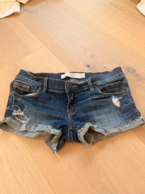 Blå vintage lågmidjade jeansshorts med slitningar - Säljer ett par blå jeansshorts med råa kanter och slitna detaljer. Shortsen har fem fickor, bälteshällor och knappgylf. Perfekta för sommaren och ger en avslappnad vibe till din outfit. Lite sönder där bak men inget som inte går att fixa!