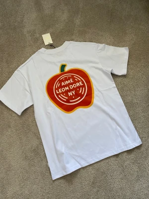 Vit Aimé Leon Dore t-shirt med tryck - Vit t-shirt från Aimé Leon Dore med stort färgglatt äpple-tryck och logga på ryggen samt mindre logga på bröstet. Klassisk passform och rund hals. Perfekt för dig som gillar streetwear och vill sticka ut lite extra.