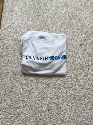 Vit stickad tröja Calvin Klein Jeans - Snygg vit stickad tröja från Calvin Klein Jeans med svart och blå logga framtill. Tröjan har rund halsringning, långa ärmar och ribbade muddar vid ärmslut och nederkant. Perfekt för en clean och stilren look.
