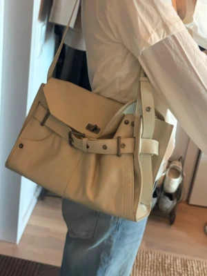 Beige axelremsväska i skinn - Snygg beige handväska i mjuk skinnimitation med axelrem som kan justeras i olika längder och dekorativt spänne framtill. Väskan har ett stort huvudfack och flera mindre innerfickor för bra organisering. Helt oanvänd i toppskick. Säljer då jag har för många väskor💖💖Köptes i butiken Urban Revovi i  Shanghai förra året