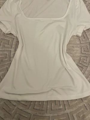 Vit topp med fyrkantig ringning - Säljer en enkel och stilren vit topp med korta ärmar och fyrkantig ringning. Toppen har en figurnära passform och är tillverkad i mjukt bomullsmaterial. Perfekt att matcha med jeans eller kjol för en clean look.