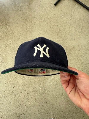 Mörkblå New Era Yankees keps - Säljer en klassisk mörkblå New Era 59Fifty keps med New York Yankees-logga broderad i vitt framtill. Kepsen har rak skärm med grön undersida och är tillverkad i ullblandning. Perfekt för dig som gillar streetwear och sportig stil. Detta är en collaboration med Essentials Fear of God vilket även står på en lädertag på baksidan. Inte ny, men utmärkt skick, inte blivit använd eftersom den inte riktigt passade mig. 